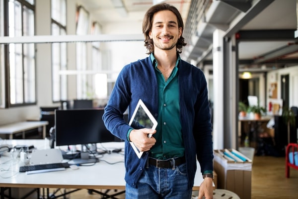 An employee stands whole holding a computer, smiling in a modern work environment.