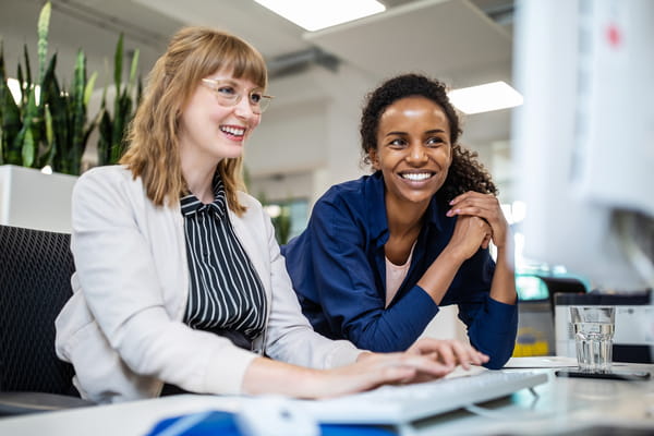 Two employees at a desk, looking at a computer screen together in a professional work setting.